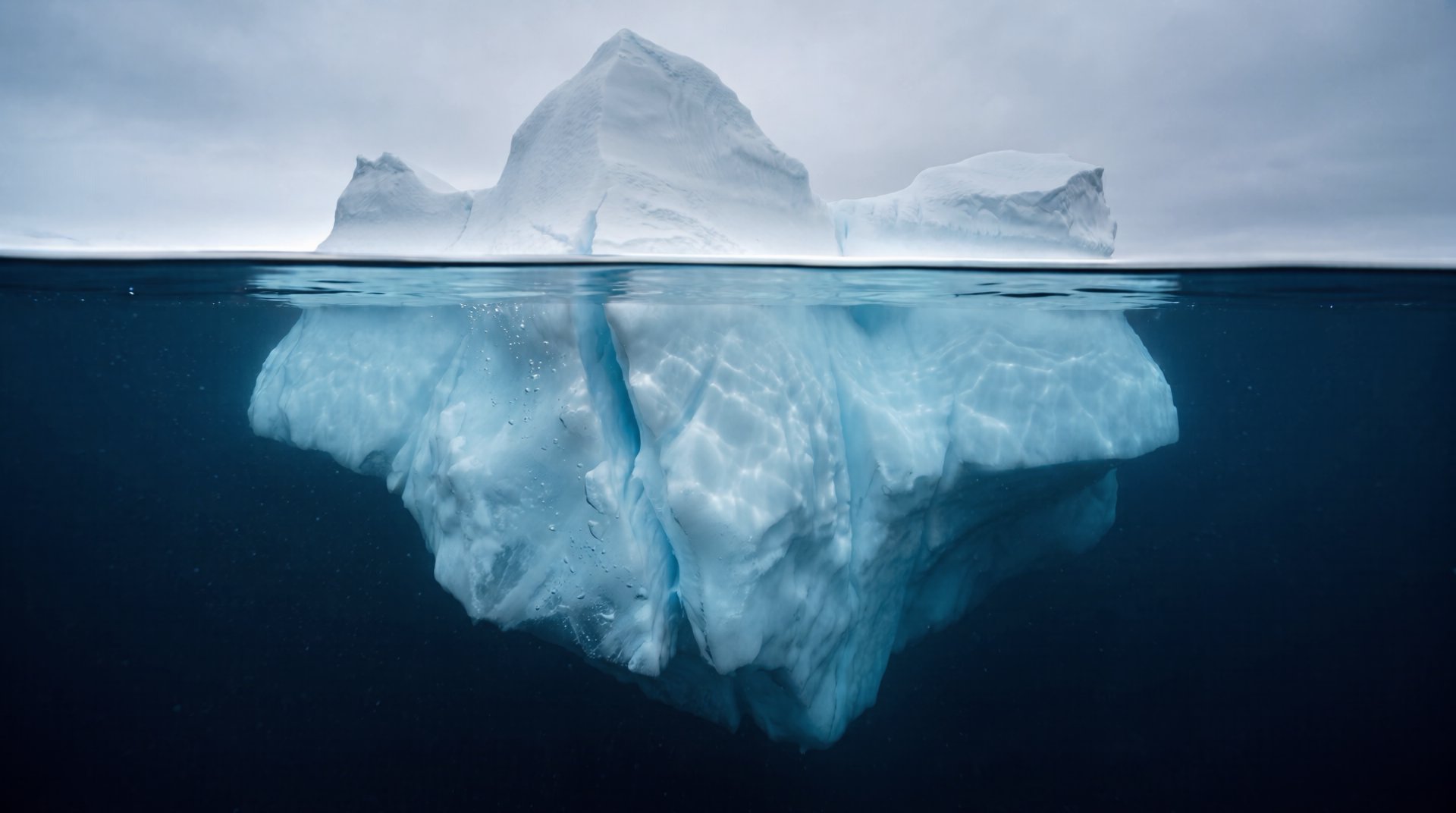 An iceberg in open polar ocean, seen above and below a straight waterline; the submerged mass is far larger than the summit.